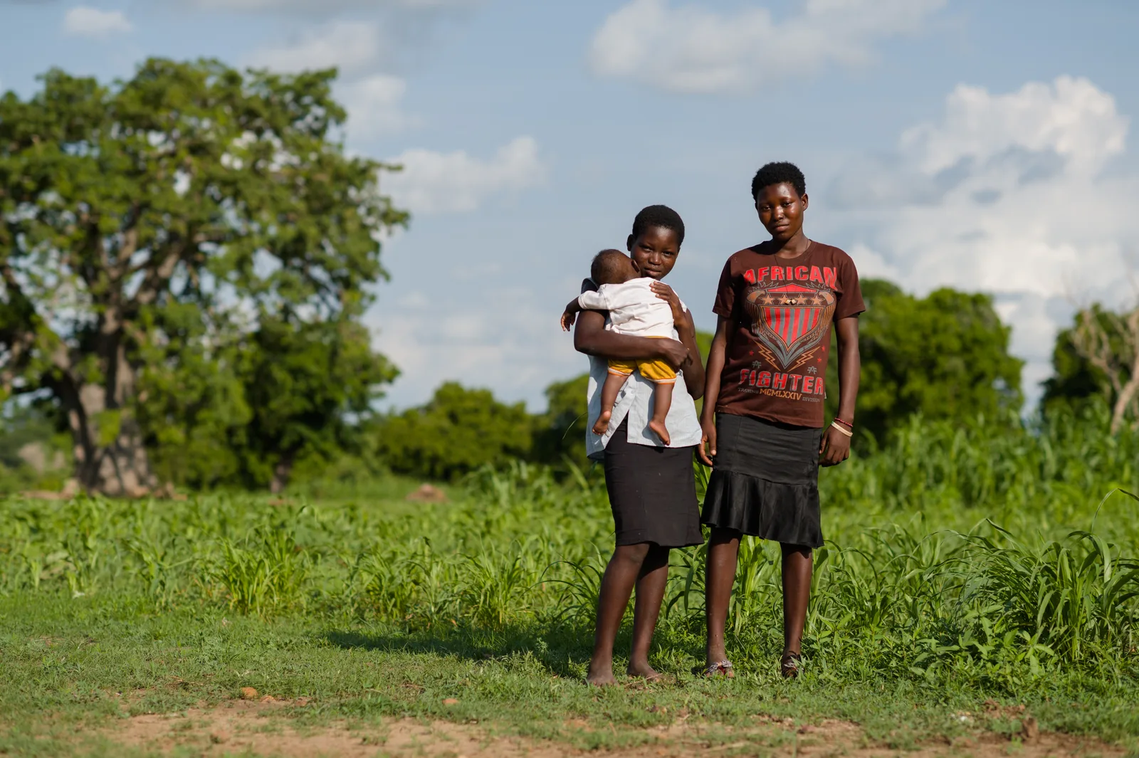 A young couple stand together outdoors, one holding a baby.