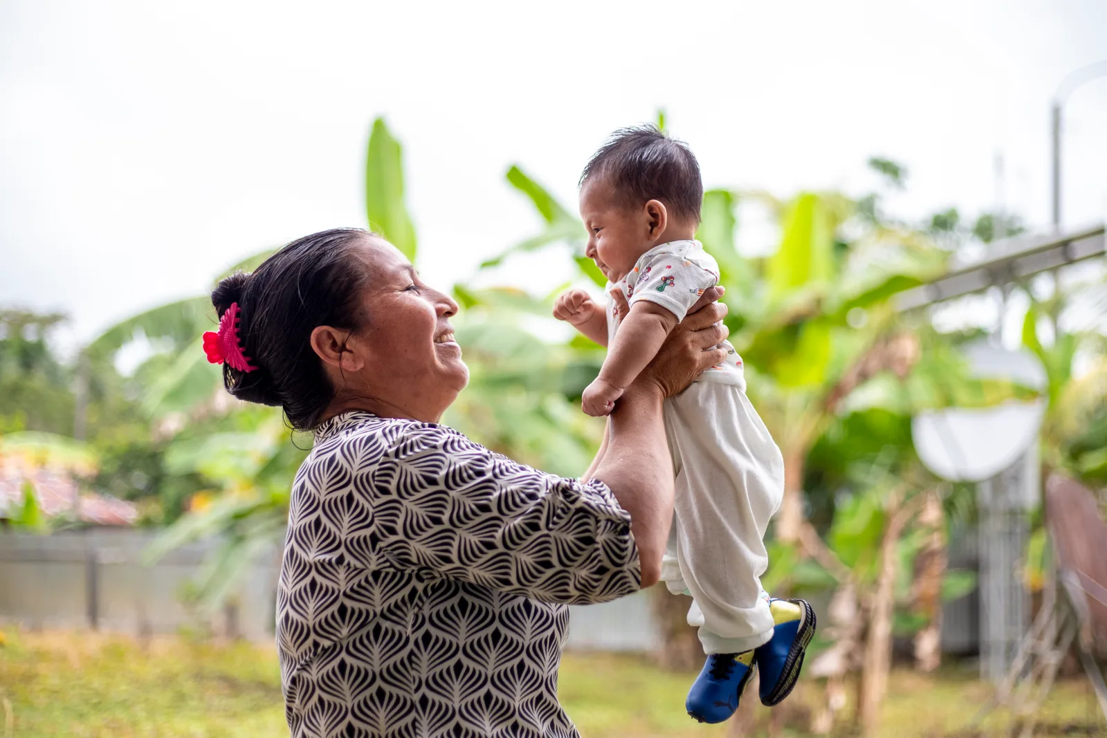 A woman smiles as she holds her baby up in front of her, outdoors.